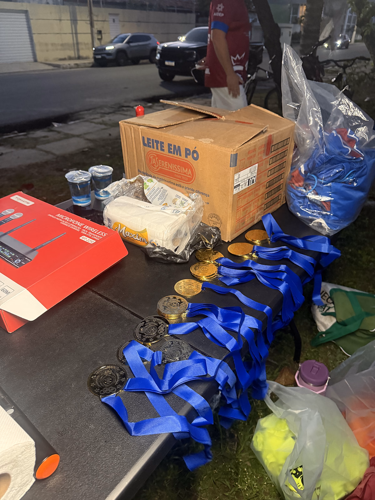 Medalhas de fita azul sobre a mesa, ao lado das mochilas e da caixa de leite em pó, antes da entrega na Copinha.