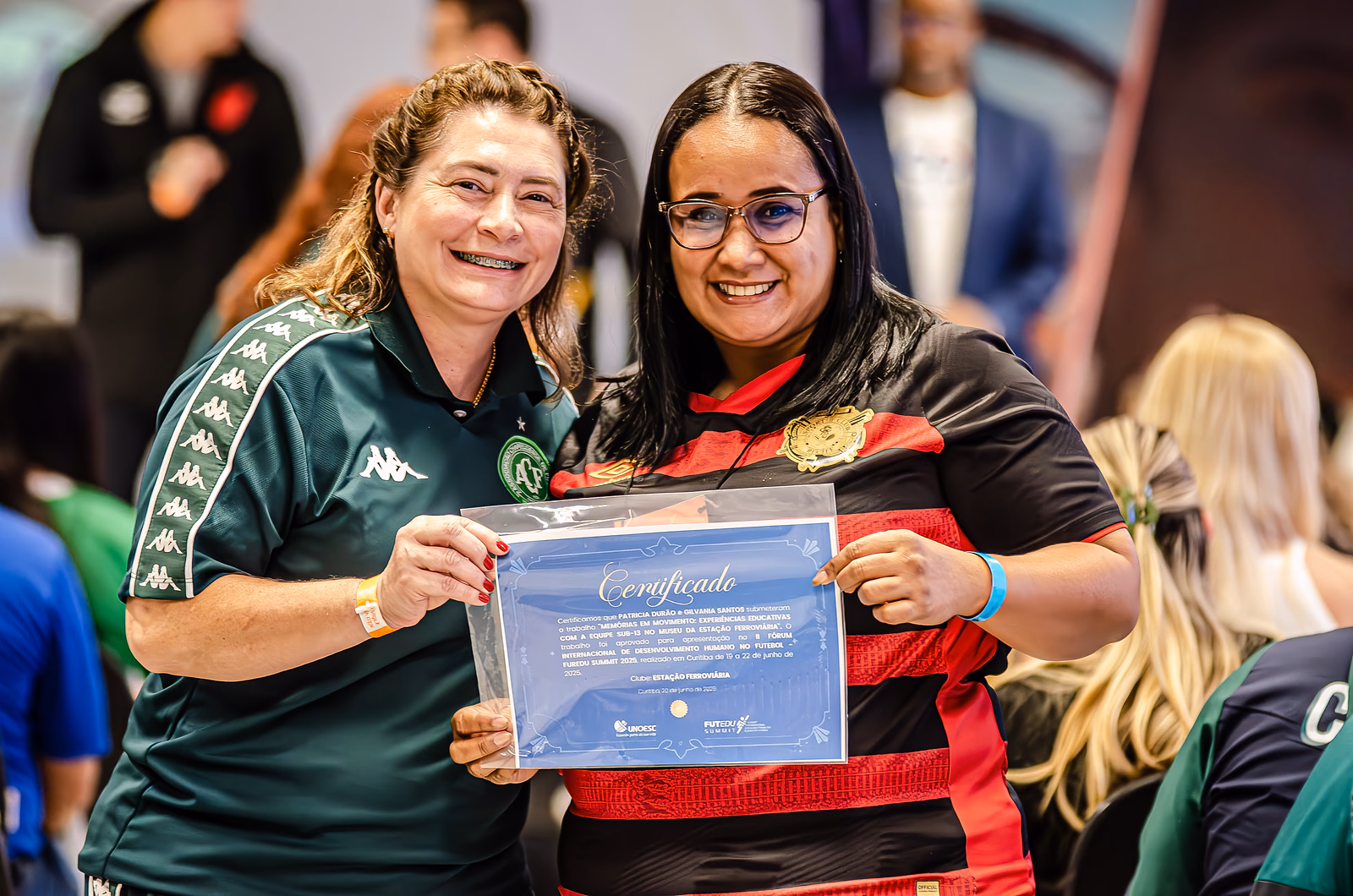 Duas educadoras seguram juntas o certificado recebido no Summit, de uniforme dos seus clubes.