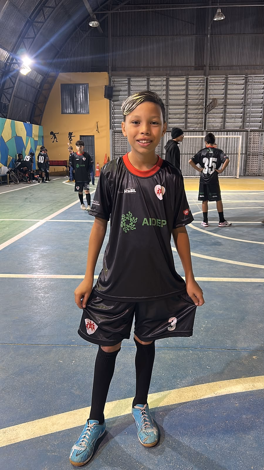 Criança de uniforme completo do projeto na quadra coberta, com a turma jogando ao fundo.