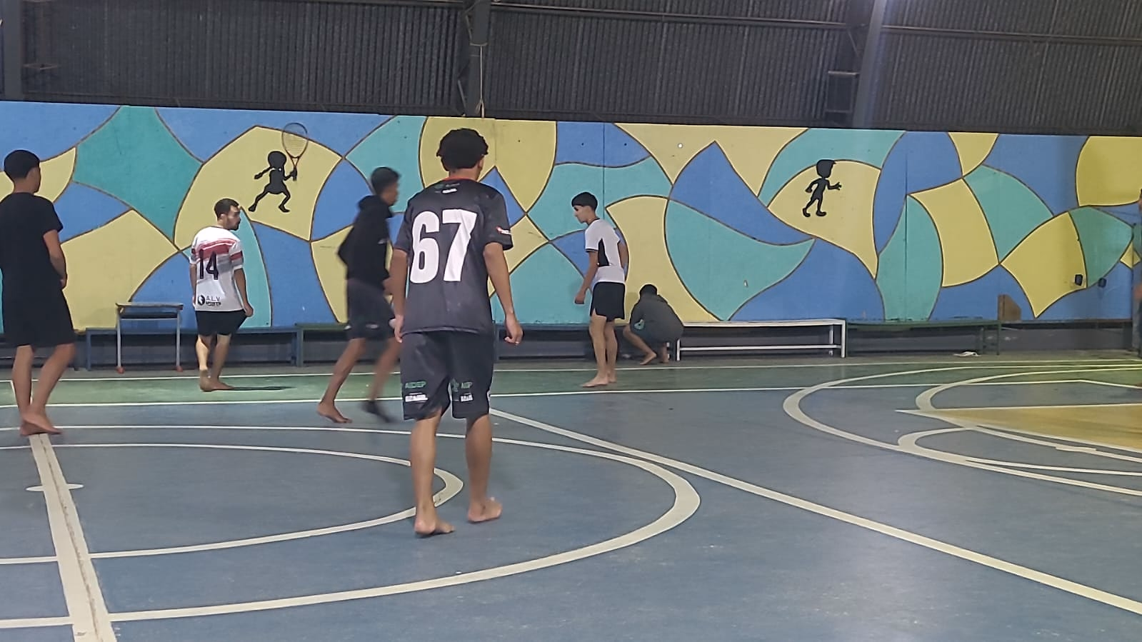 Adolescentes disputam a bola junto ao mural da quadra, durante a atividade.