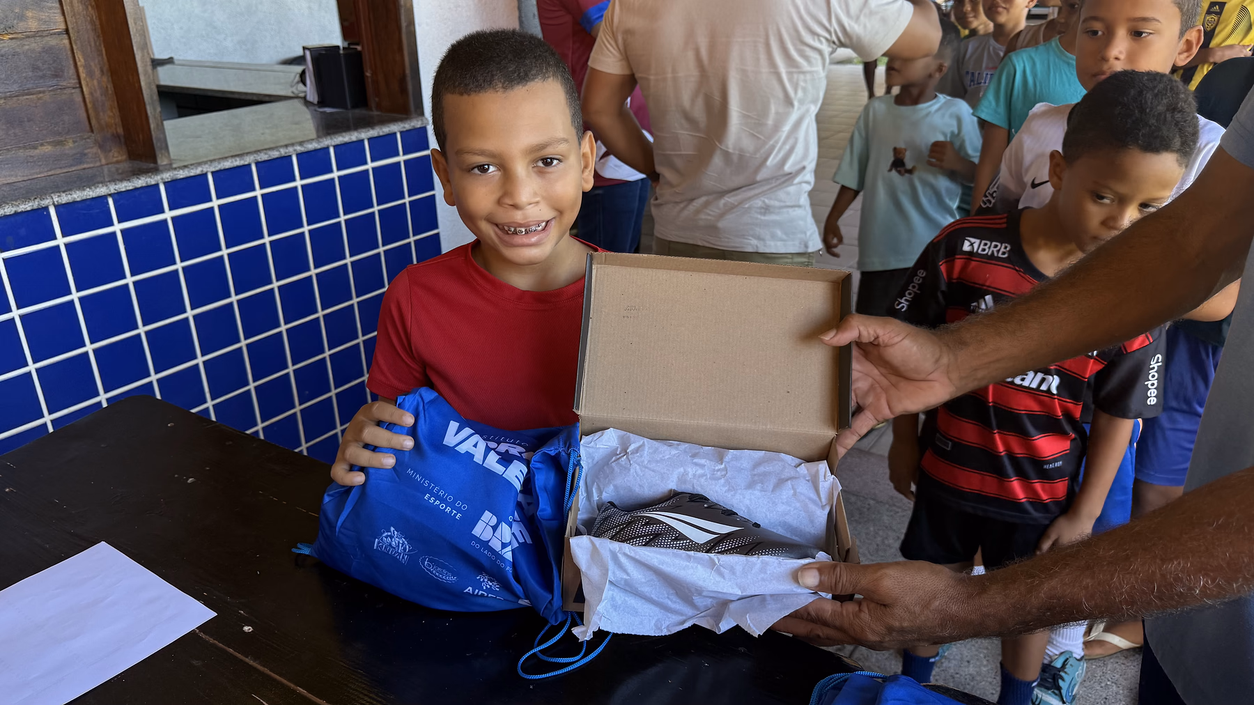 Menino sorri ao abrir o kit entregue pelo projeto, com uniforme e chuteiras.