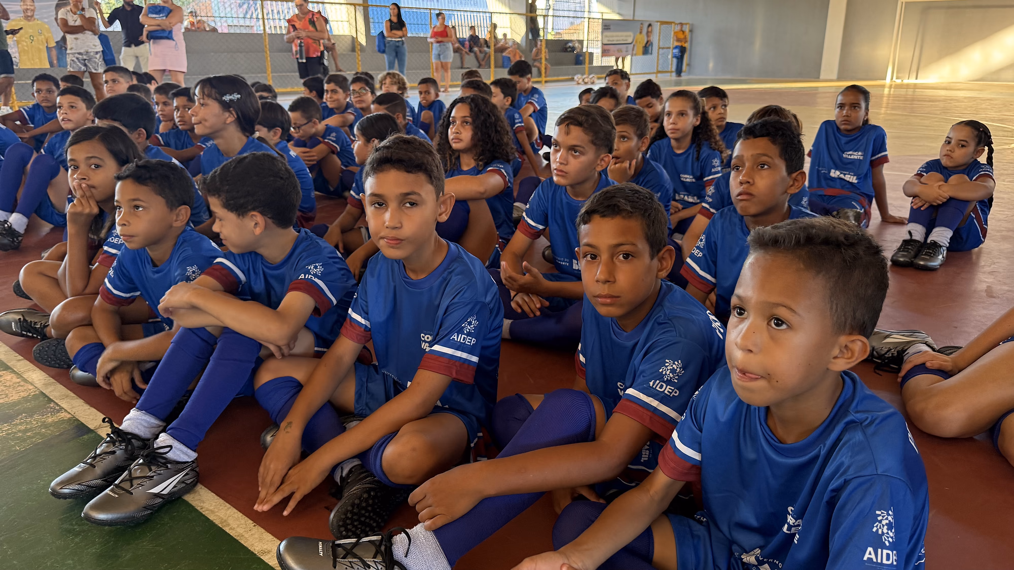 Meninas e meninos sentados na quadra do ginásio, atentos à abertura do polo de Poço Verde.