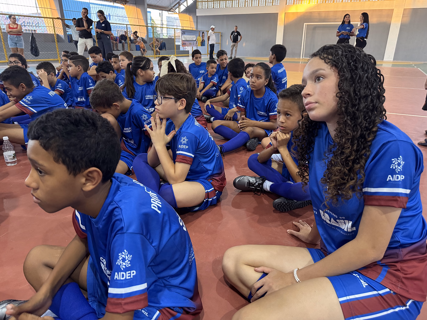 Crianças de uniforme sentadas no chão do ginásio de Poço Verde, acompanhando a abertura de perto.